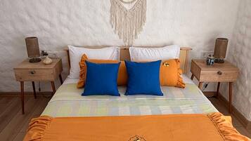 A bed with an orange blanket, two blue pillows, and a macrame wall hanging.