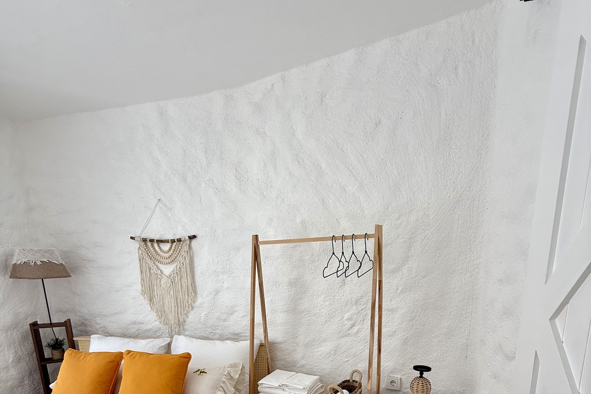 Un dormitorio con cama, mesita de noche, lámpara, un colgador de macramé en la pared y un perchero.