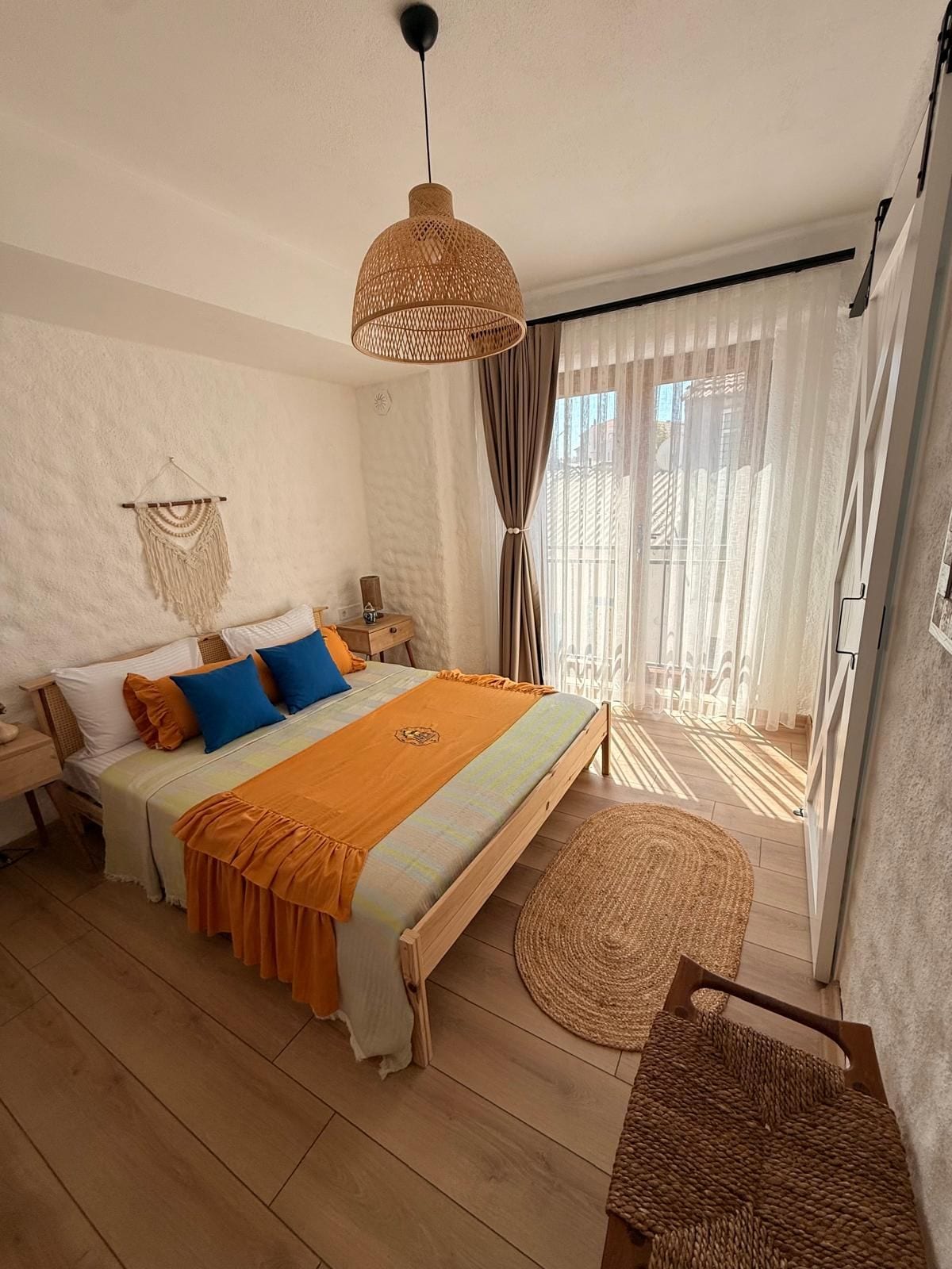 A bedroom with a bed, bedside table, lamp, curtains, and a view of the outdoors.
