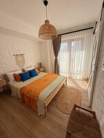 A bedroom with a bed, bedside table, lamp, curtains, and a view of the outdoors.