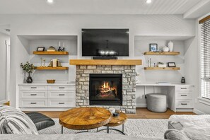 A modern living room with a stone fireplace, white furniture, and a flat-screen TV mounted above the fireplace. - The Bluebird Suite - Mountain Views - Hot Tub - 2BR (Canmore)