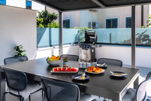 An outdoor patio with a table set for dining, a grill, and a view of the building in the background. - Holiday home Val (Šibenik)