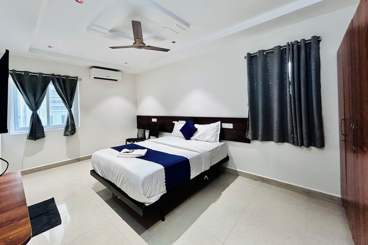 A hotel room with a bed, a ceiling fan, a window with curtains, and a door.