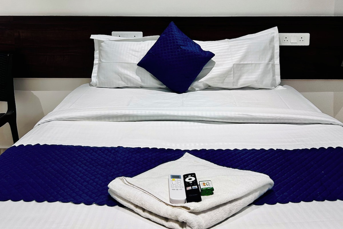 A neatly made bed with white linens, a blue pillow, and a folded towel with a remote control and a card on it.
