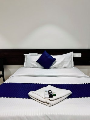A neatly made bed with white linens, a blue pillow, and a folded towel with a remote control and a card on it. - Orion Signature Hotel  (Hyderabad)