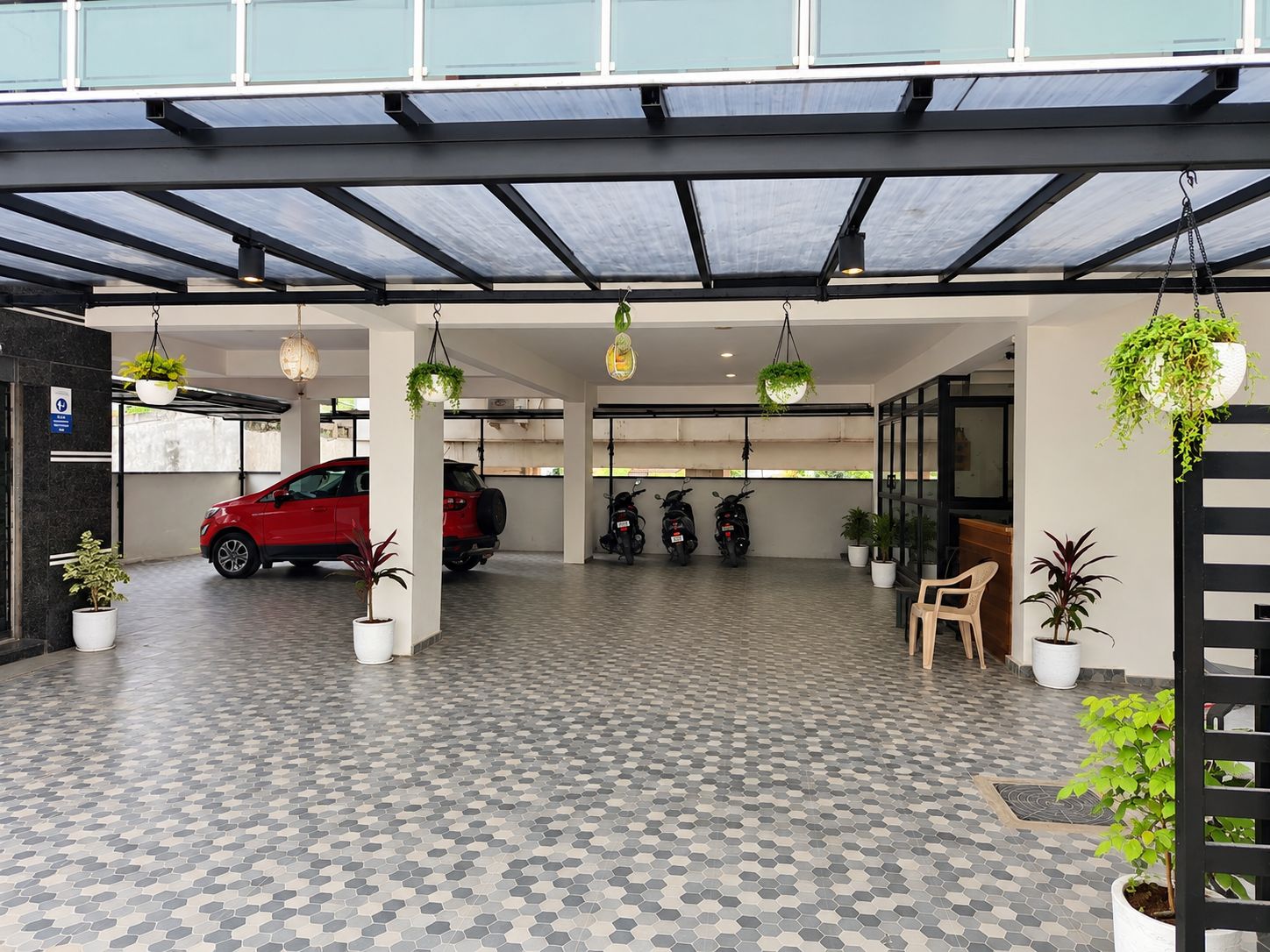 Área de estacionamento coberta com um carro vermelho e algumas scooters estacionadas, plantas em vasos e uma área de descanso com uma cadeira.