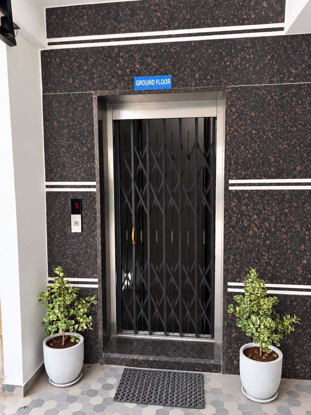 Ein Gebäudeeingang mit einem Schild 'GROUND FLOOR', einem Metallgitter, einer digitalen Aufzugsanzeige und zwei Pflanzkübeln.
