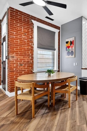 Una habitación con pared de ladrillo, una mesa redonda de madera con sillas, un ventilador de techo y una ventana con persianas.