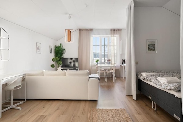 A modern, well-lit living room with a white sofa, a dining area with a table and chairs, and a bed with a patterned bedspread.