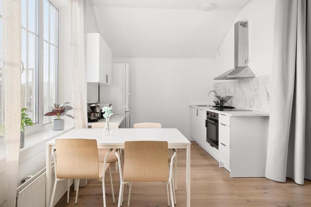 Une cuisine moderne avec un plan de travail blanc, une cuisinière et un évier. On y trouve une table à manger avec des chaises et une fenêtre avec des rideaux.