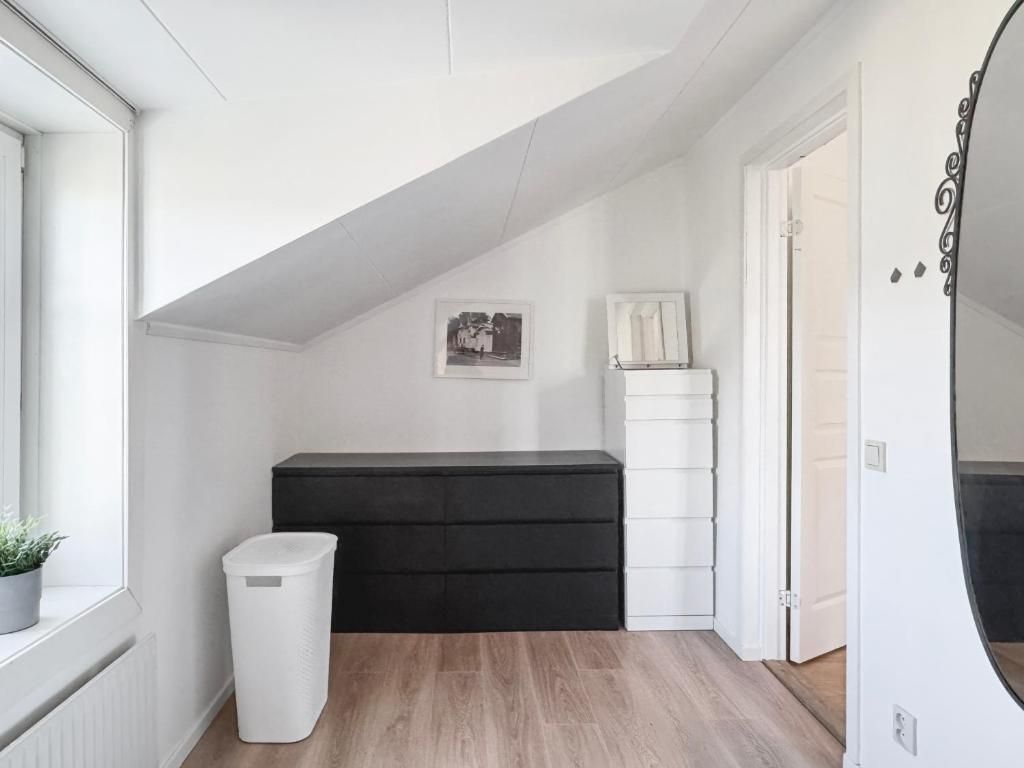 Quarto com teto inclinado, um móvel de armazenamento preto, uma lixeira branca e uma porta branca.