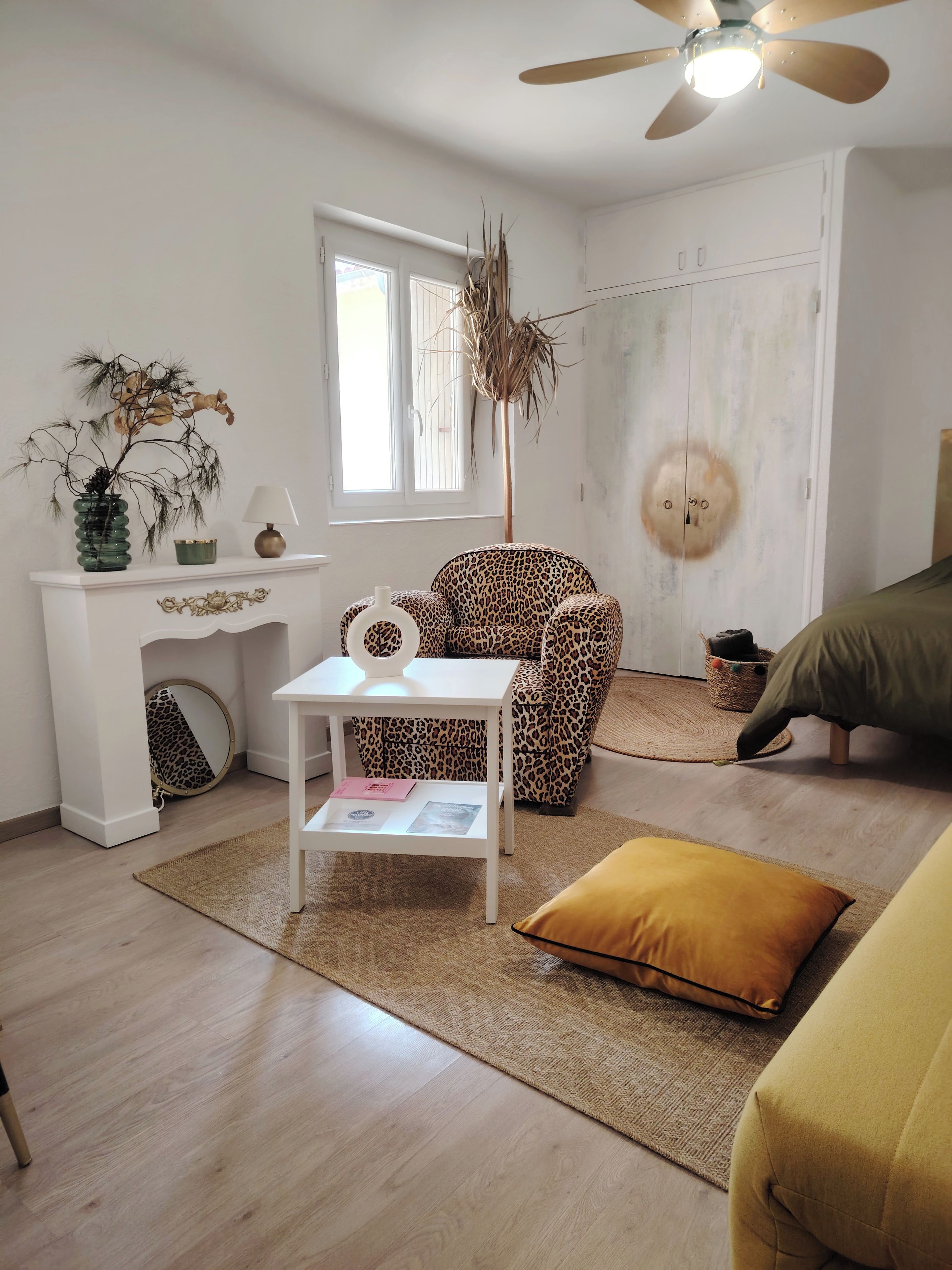 A room with a white sofa, a leopard print chair, a small white table with a lamp, a decorative plant, and a ceiling fan.
