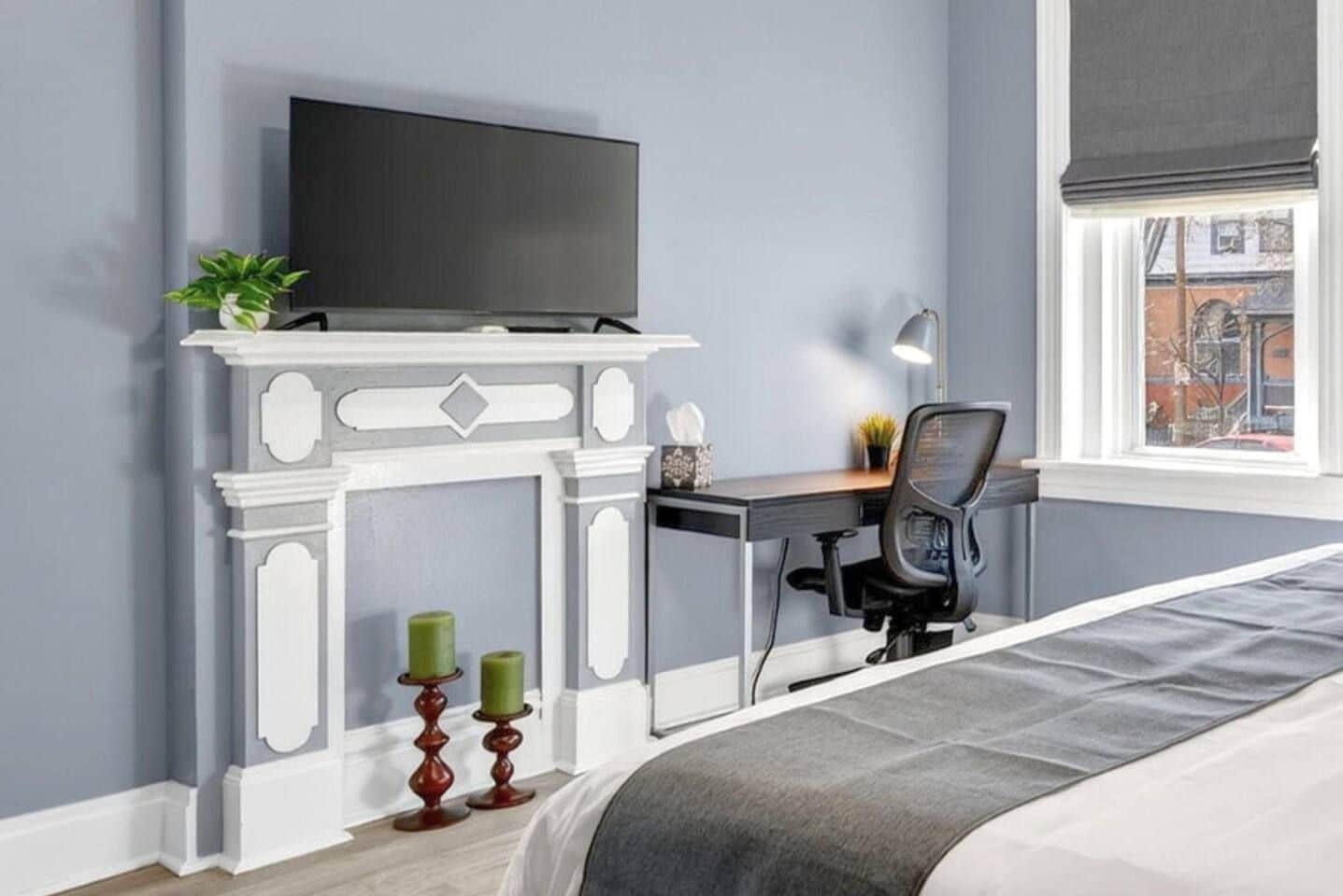 Een slaapkamer met een bed, een bureau met stoel, een televisie boven een open haard, en een raam met uitzicht op gebouwen.