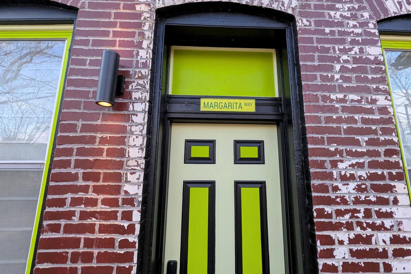 Een gevel van een gebouw met een bakstenen buitenkant, een deur in groen en zwart, en een bord boven de deur met de tekst 'MARGARITA WAY'.