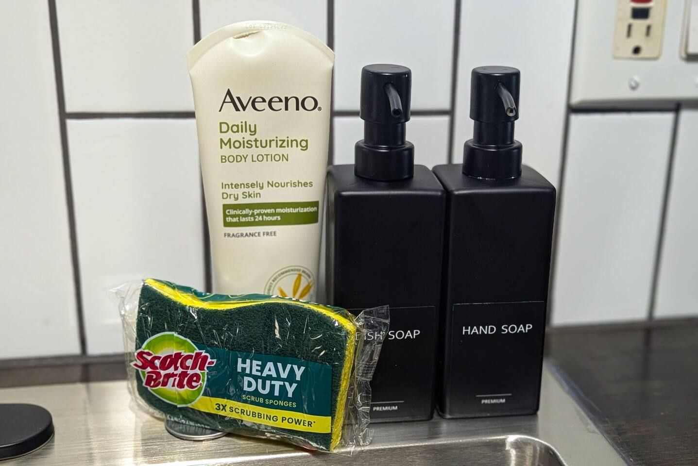 Ein Badezimmer-Waschtisch mit Aveeno Daily Moisturizing Body Lotion, Scotch-Brite Heavy Duty Schwämmchen und zwei Seifenspendern.