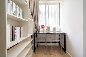 A room with a desk, two chairs, a bookshelf, and a window with sheer curtains.