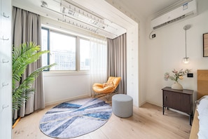 A modern bedroom with a large window, a round rug, a wooden floor, a small round ottoman, a nightstand with a vase of flowers, and a hanging light fixture.