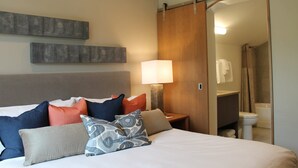 Une chambre d’hôtel comprenant un lit, des oreillers, une table de chevet, une lampe, une porte coulissante donnant accès à la salle de bain et une œuvre d’art accrochée au mur.