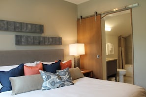 Quarto de hotel com cama, almofadas, mesa de cabeceira, luminária, porta de correr para um banheiro e uma obra de arte na parede.
