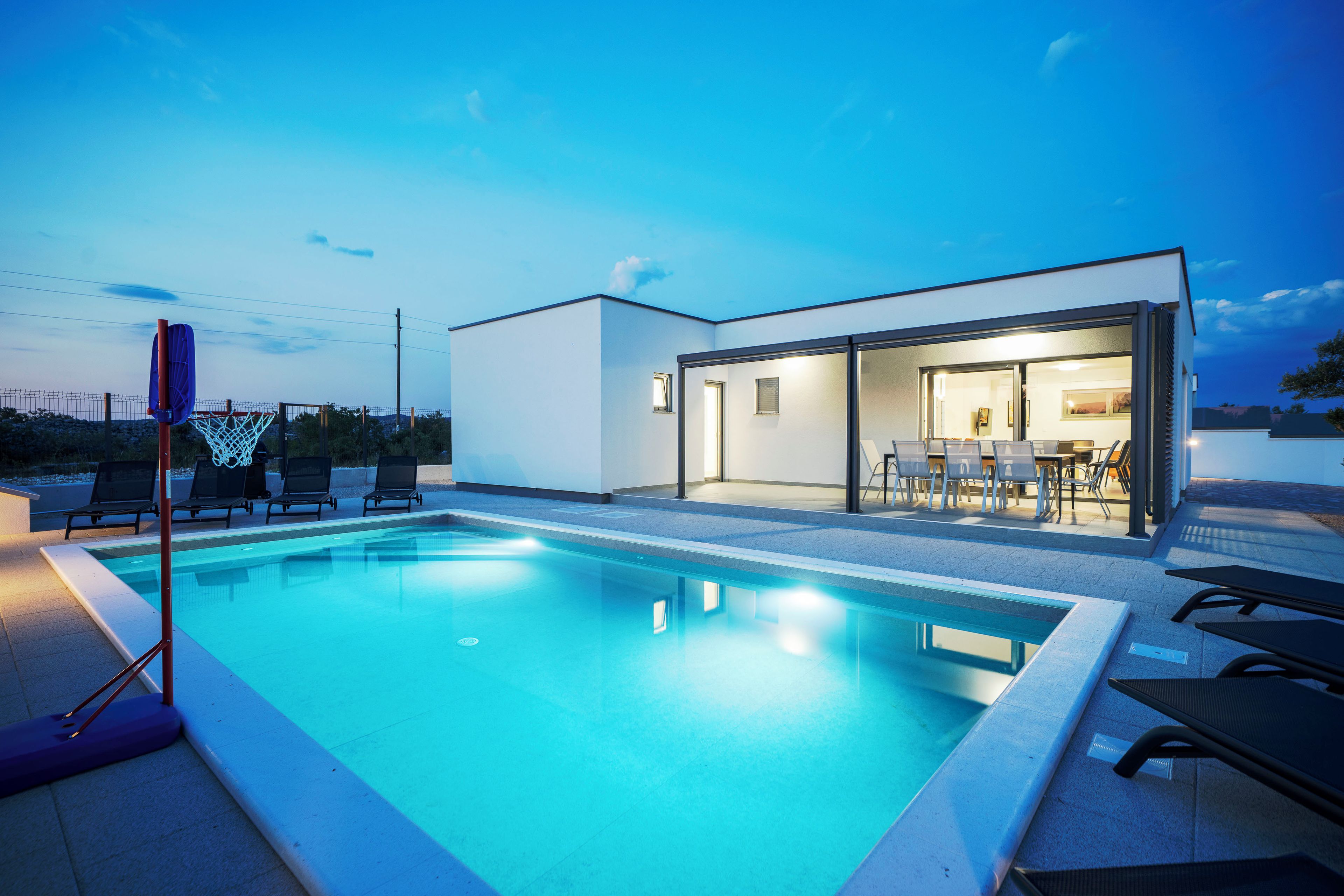 Un espace piscine extérieur moderne, avec un ciel dégagé, un panier de basketball et un bâtiment doté de portes vitrées.