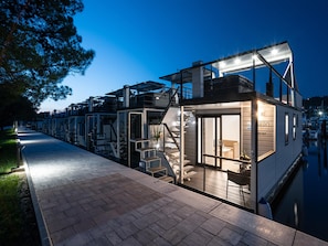 A waterfront property with a glass-enclosed structure, outdoor seating, and a staircase leading to the upper level.