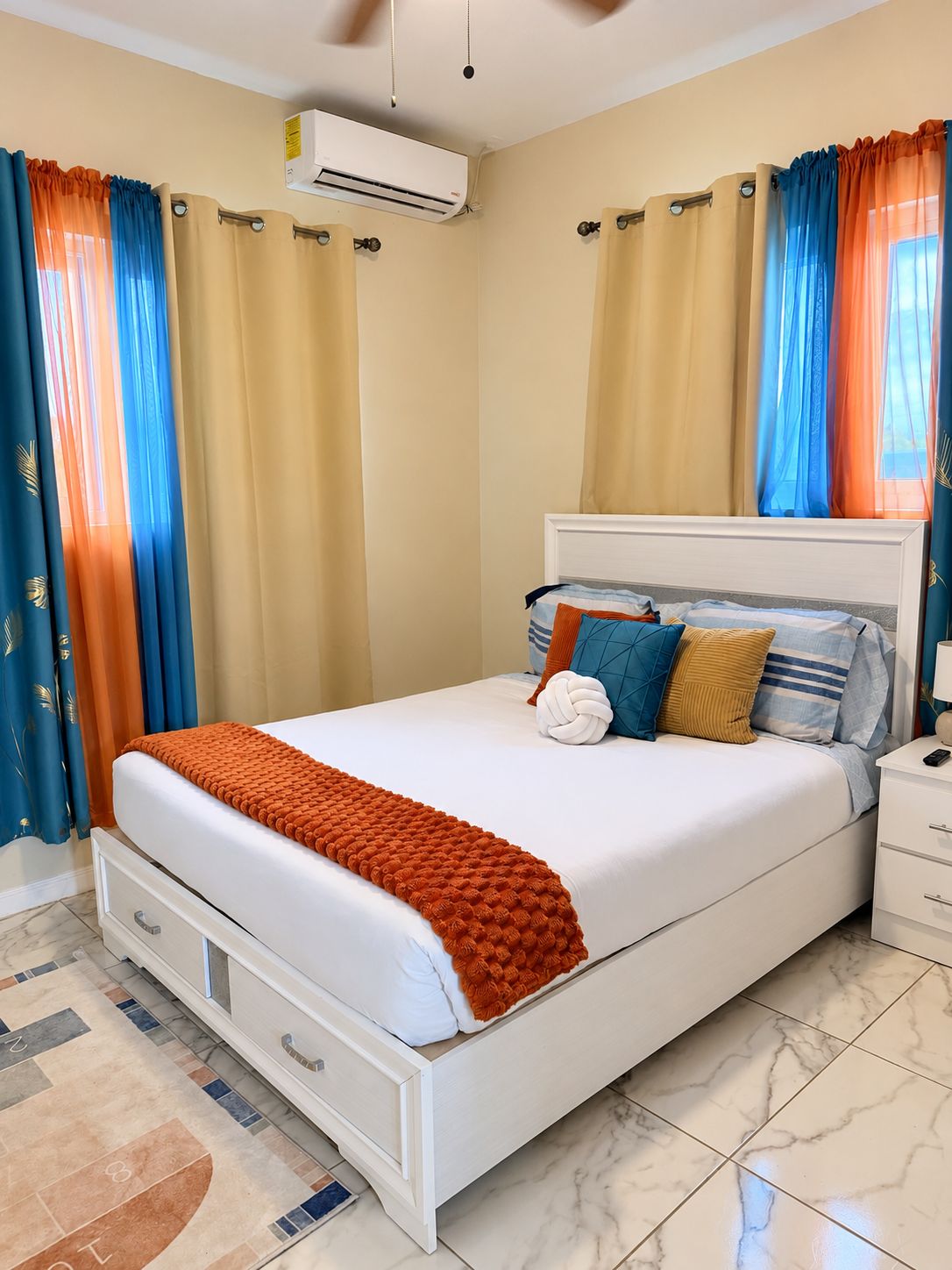 A hotel room with a bed, a nightstand, a drawer, an air conditioning unit, and curtains.