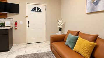 A living room with a brown leather sofa, a white door with a window, a small white table with a vase of flowers, and a mounted flat-screen TV.