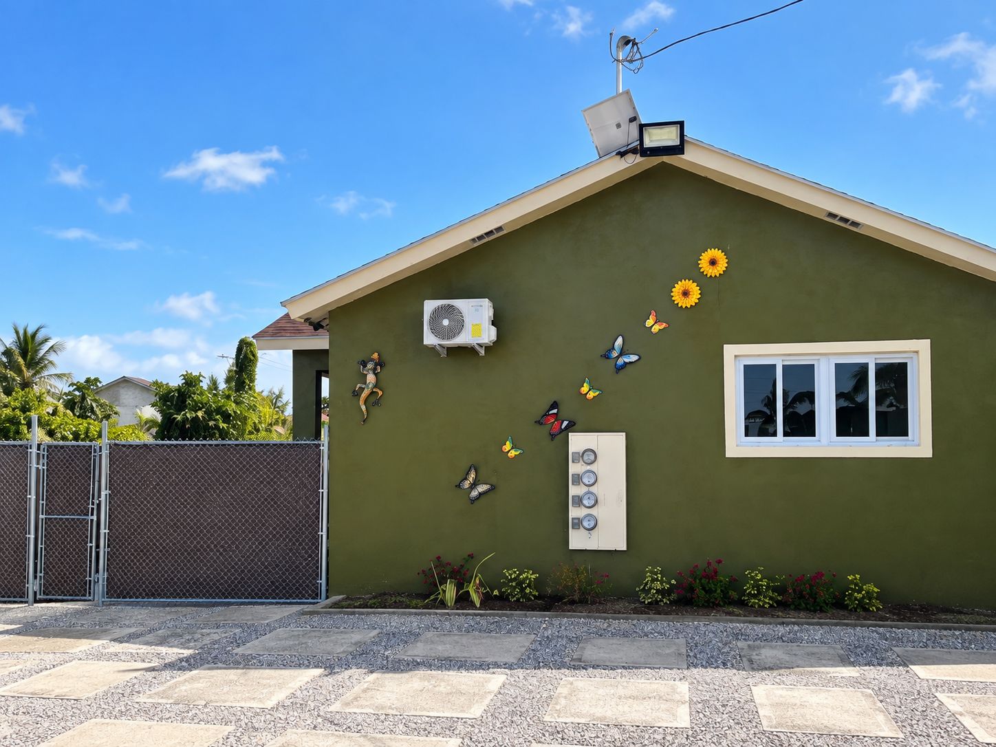 A house with a green exterior, a solar panel on the roof, an air conditioning unit, a window with a view, and decorative butterflies and flowers on the wall.