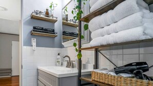 A bathroom with a white sink, a mirror, a towel rack with white towels, a shelf with makeup items, and a plant. - Charming Kirkwood Two-Bedroom (St. Louis)