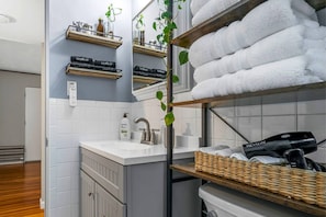 Un baño con un lavamanos blanco, un espejo, un perchero con toallas blancas, un estante con artículos de maquillaje y una planta.