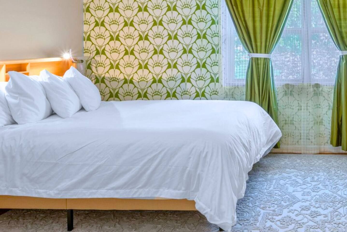 Una cama con ropa de cama blanca, un papel tapiz estampado y una ventana con cortinas verdes.