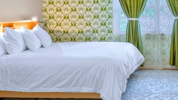 Una cama con ropa de cama blanca, un papel tapiz estampado y una ventana con cortinas verdes.