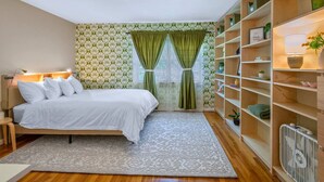 A bedroom with a bed, a nightstand, a bedside lamp, a window with curtains, a bookshelf, and a radiator. - Charming Kirkwood Two-Bedroom (St. Louis)