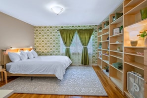 Un dormitorio con cama, mesita de noche, lámpara de pie, ventana con cortinas, estantería y radiador.