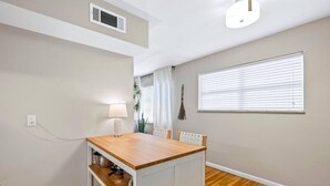 A room with a wooden table, a lamp, a window with blinds, a wall-mounted air vent, and a power outlet. - Charming Kirkwood Two-Bedroom (St. Louis)