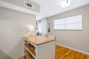 Una habitación con una mesa de madera, una lámpara, una ventana con persianas, un respiradero en la pared y un enchufe.