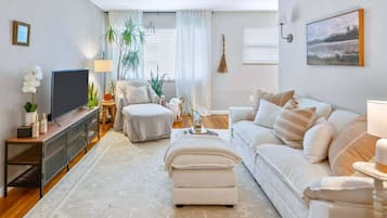 Una sala de estar con sofá, reposapiés, televisor y un ventanal con cortinas.