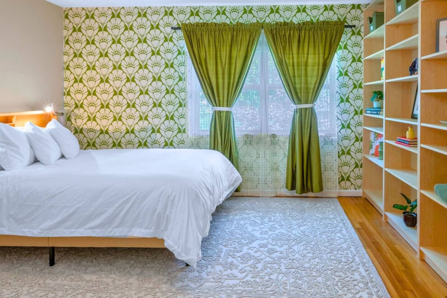A bedroom with a bed, a patterned wallpaper, a window with green curtains, and a bookshelf.