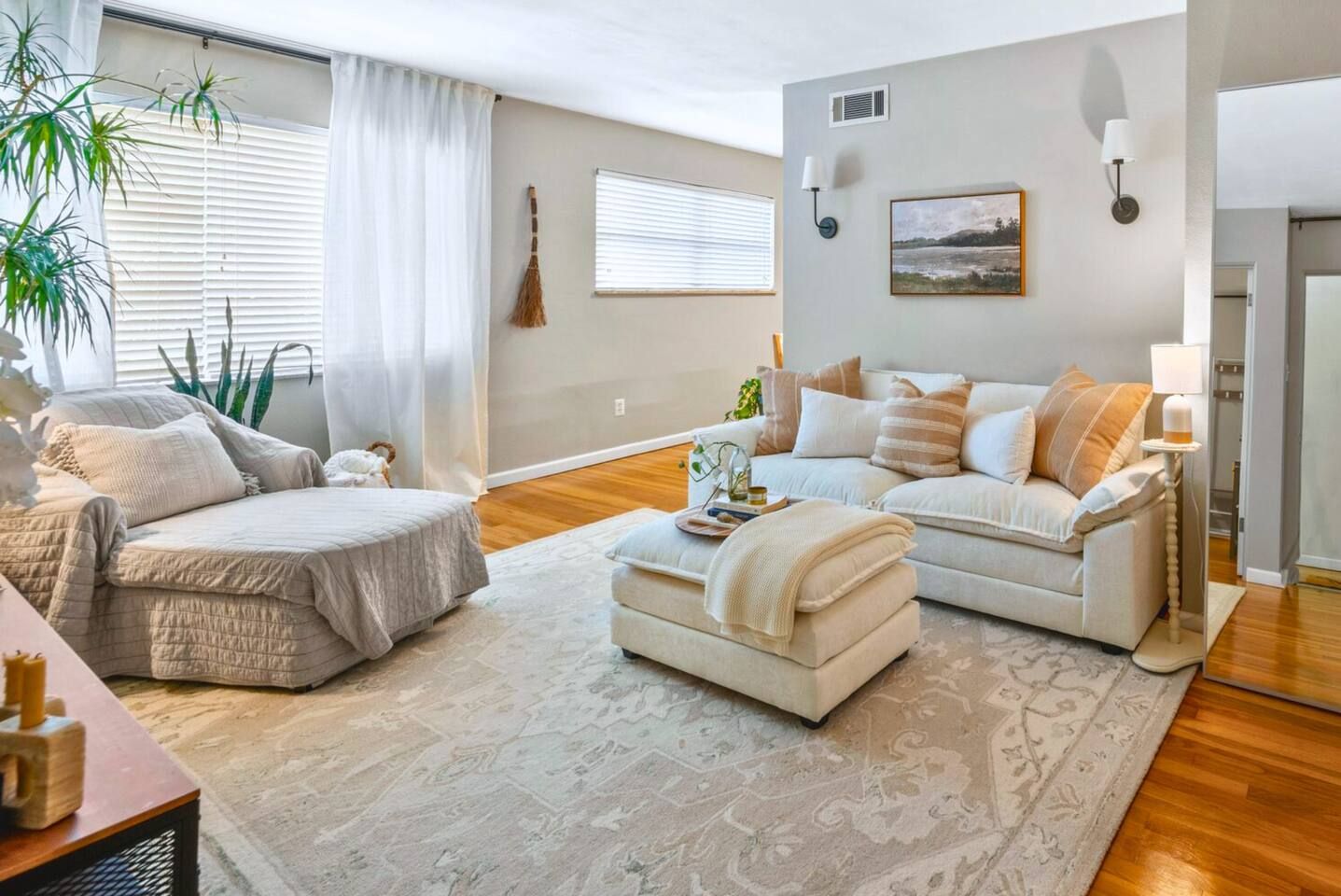 Una sala de estar con un sofá, una silla, una mesa de centro y una alfombra. Hay plantas y un cuadro en las paredes.