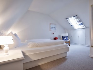 A bedroom with a sloped ceiling, a bed with white bedding, a nightstand, a chair with a star pillow, a small table with a red object, and a wardrobe. - Holiday Apartment With 1 Bedroom (Kampen)