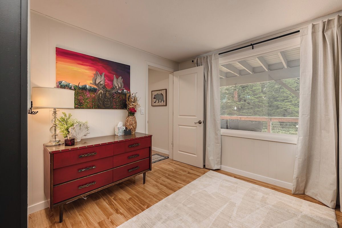 Une pièce avec un plancher en bois, une commode rouge, un tableau, une porte et une fenêtre avec des rideaux.