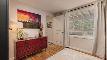 Une pièce avec un plancher en bois, une commode rouge, un tableau, une porte et une fenêtre avec des rideaux.