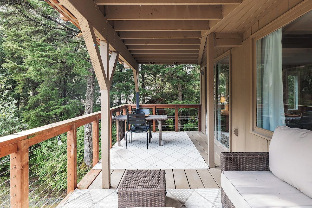 A covered outdoor area with a table and chairs, a sofa, and a view of dense forest.