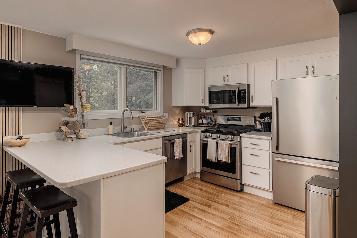 A modern kitchen with white cabinetry, stainless steel appliances, a large window, and a flat-screen TV mounted above the counter.
