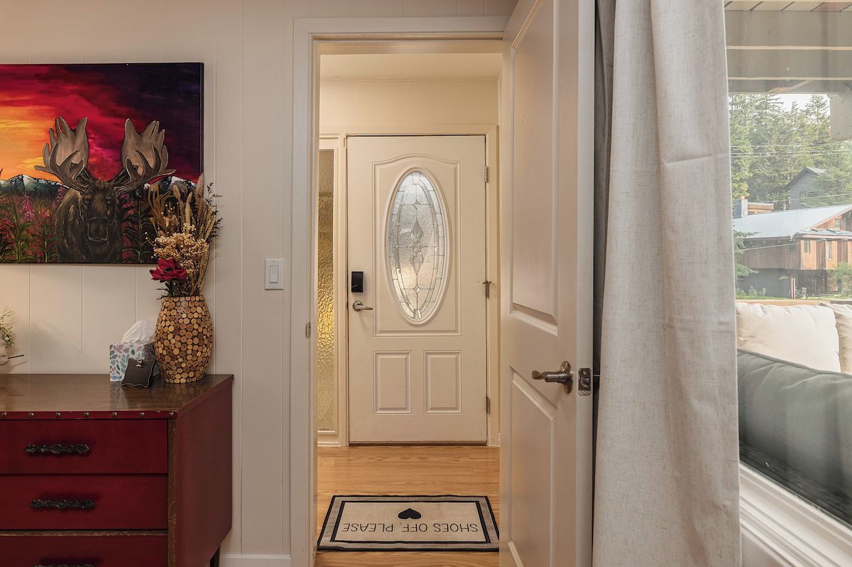 A hallway leading to a door with a frosted glass panel, a red dresser with drawers, a vase with dried flowers, and a doormat with a message.