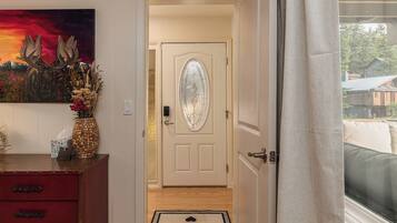A hallway leading to a door with a frosted glass panel, a red dresser with drawers, a vase with dried flowers, and a doormat with a message.