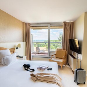 Habitación de hotel con cama, una silla, televisión, maleta y balcón con vistas.