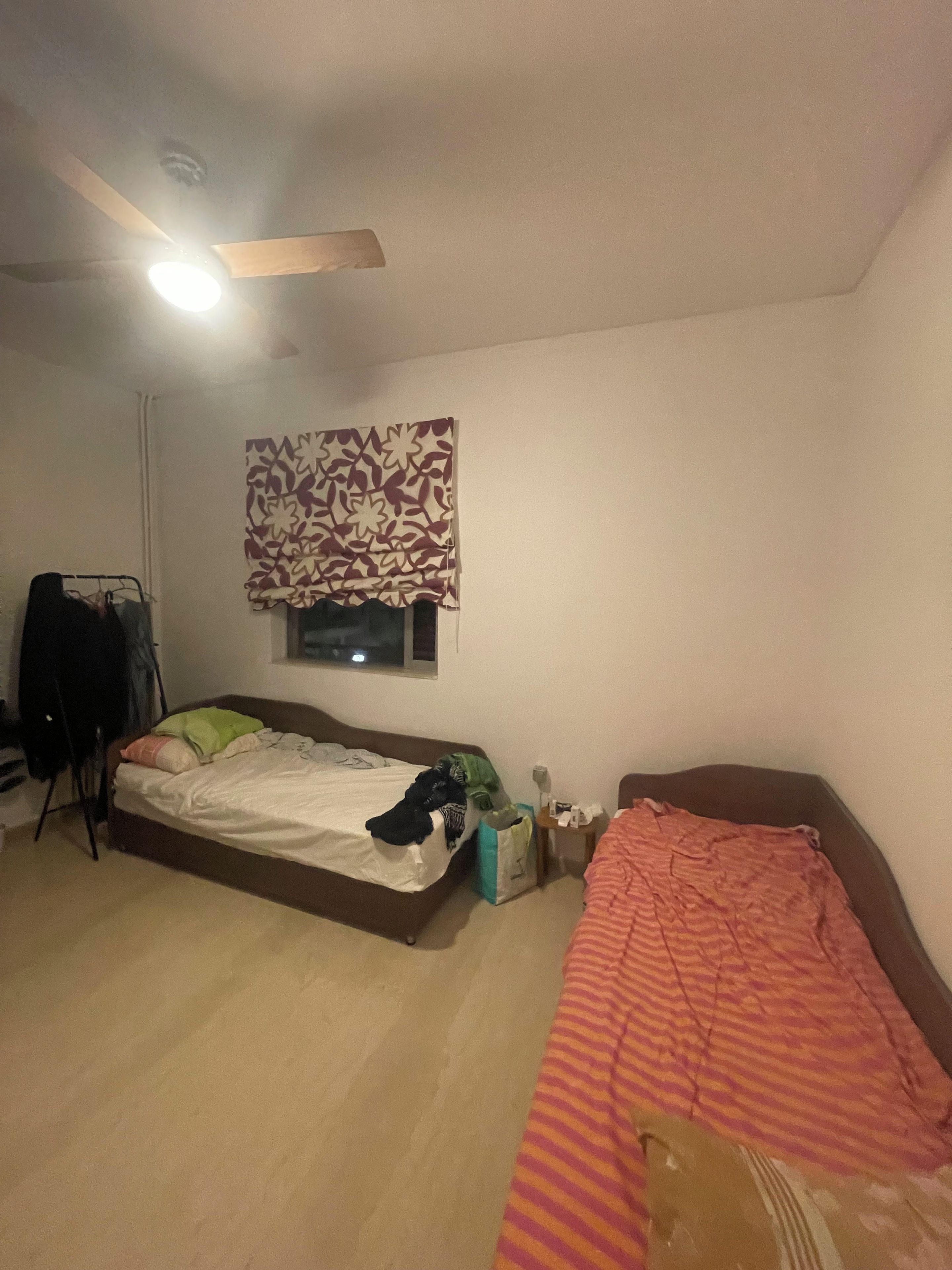 A bedroom with a bed, a bedside table, a window with patterned curtains, a ceiling fan, and a wall-mounted light fixture.