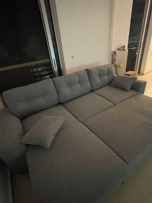 A grey sofa with a cushion, positioned against a wall with a door and a radiator.