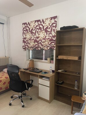 A room with a desk, chair, bookshelf, radiator, window with blinds, and a potted plant.